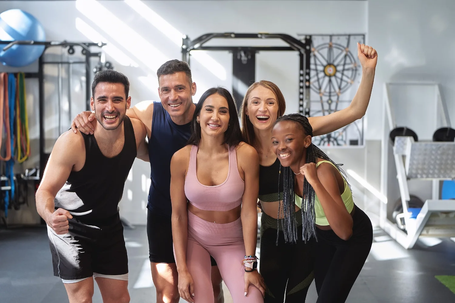 Group of five diverse gym members smiling and posing together in a fitness studio, emphasizing community and camaraderie in a gym environment.